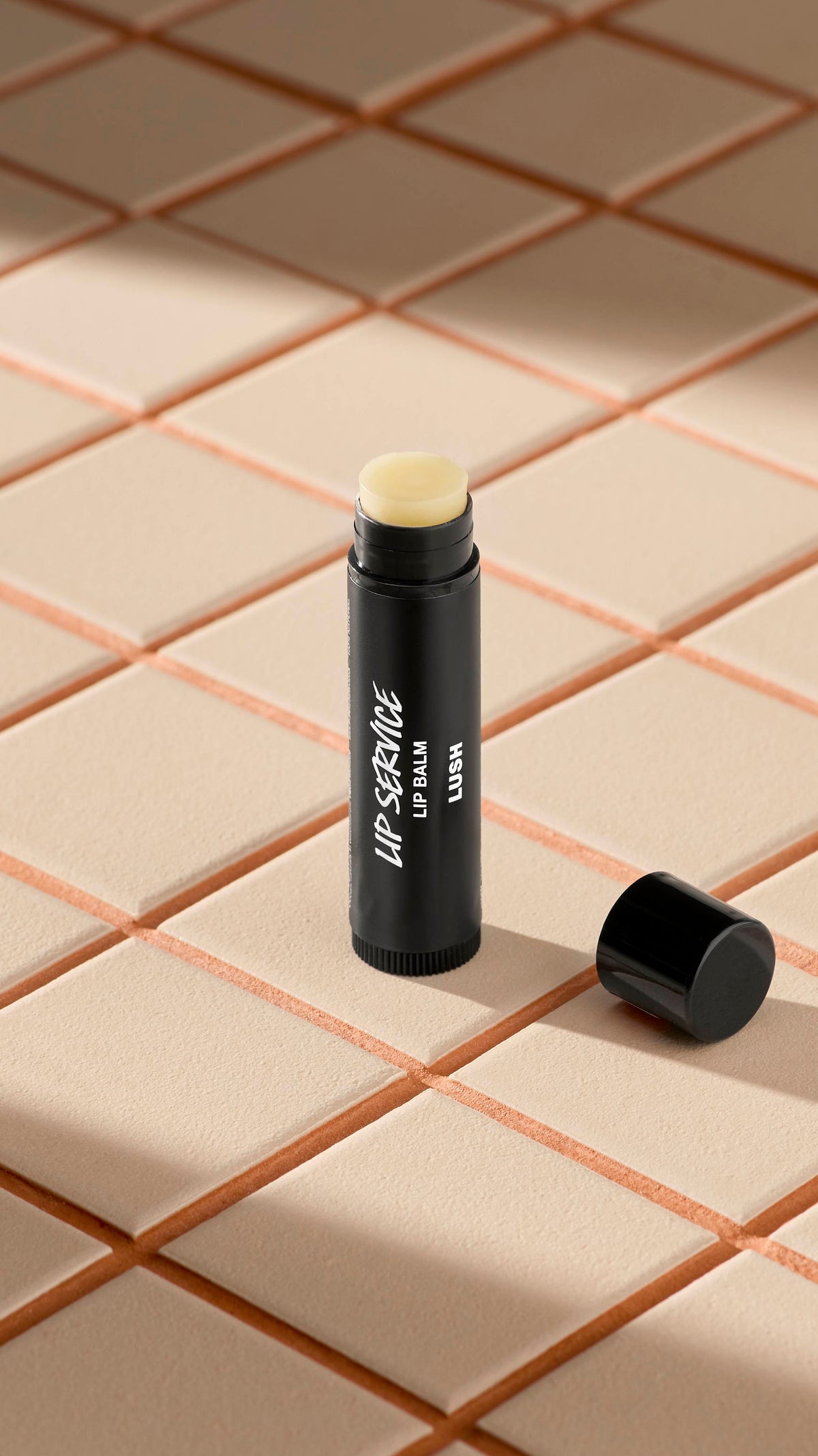 A black tube of Lush 'Lip Service' lip balm stands upright on light-coloured square tiles. The black cap is off, lying next to the base, revealing the pale, creamy balm inside. Soft shadows fall across the tiled surface.